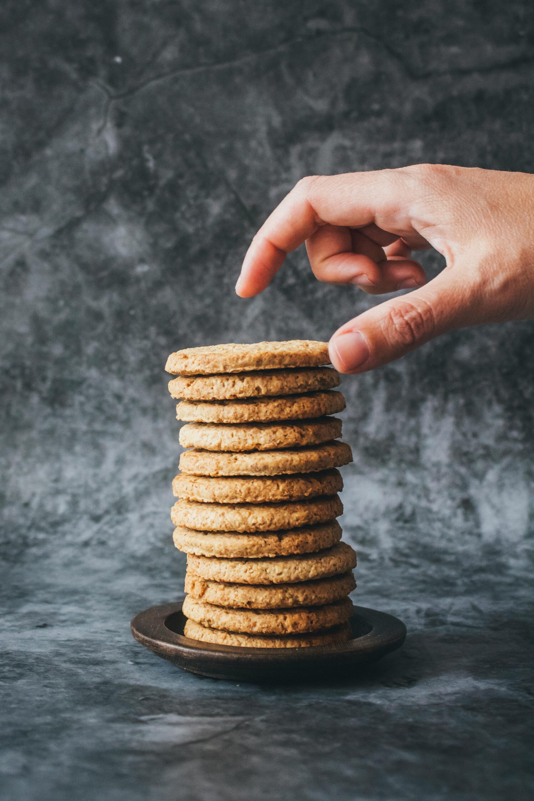 Hand reaching for delicious homemade cookies stacked on a wooden plate against a textured background.