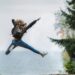 Energetic woman leaping in outdoor forest setting with lake view, showcasing freedom and joy.